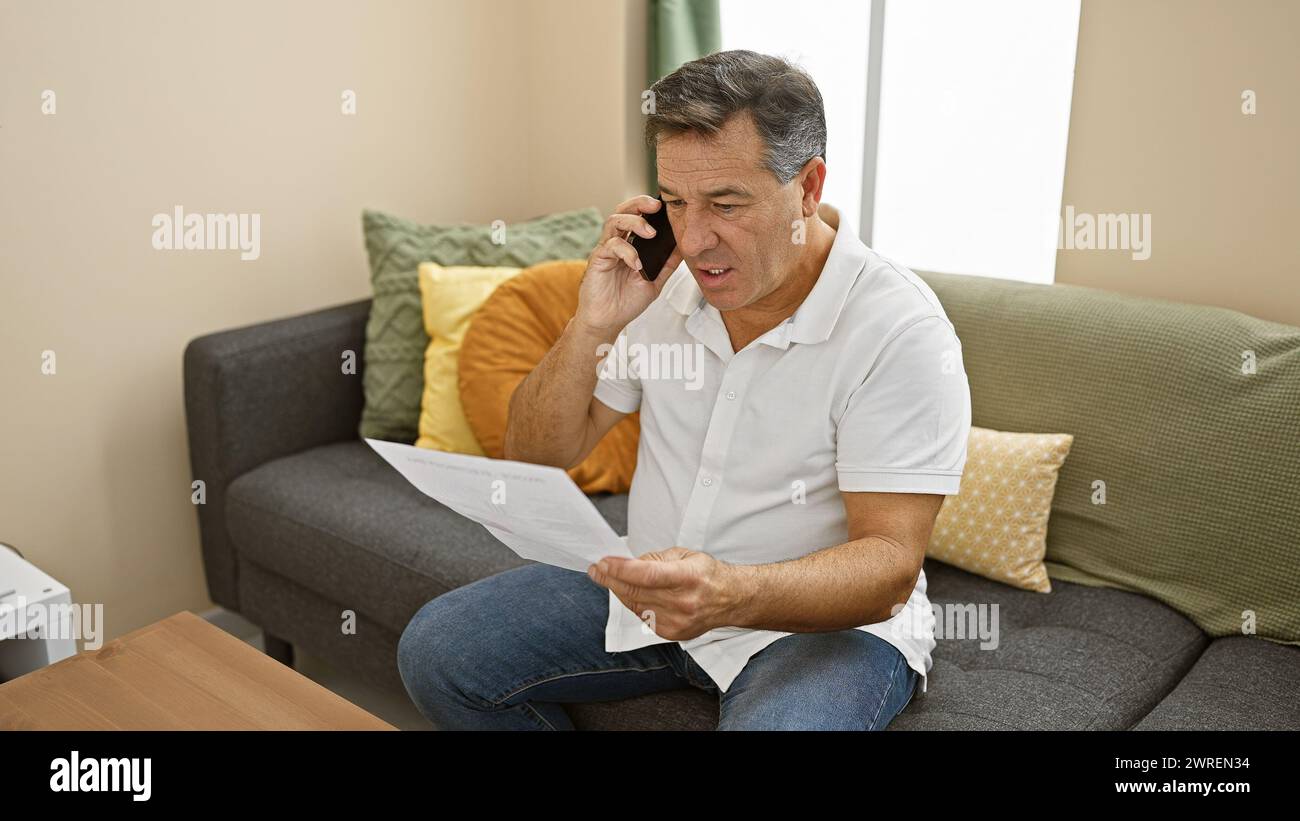 Homme d'âge moyen en chemise blanche parlant au téléphone tout en examinant le document, assis sur un canapé gris dans un intérieur de la maison Banque D'Images