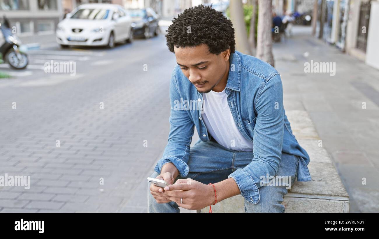 Beau jeune homme utilisant un smartphone tout en étant assis décontracté sur le trottoir de la ville. Banque D'Images