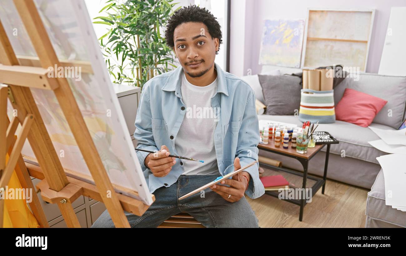 Bel homme peignant sur toile dans un studio d'art rempli de fournitures et de lumière naturelle. Banque D'Images