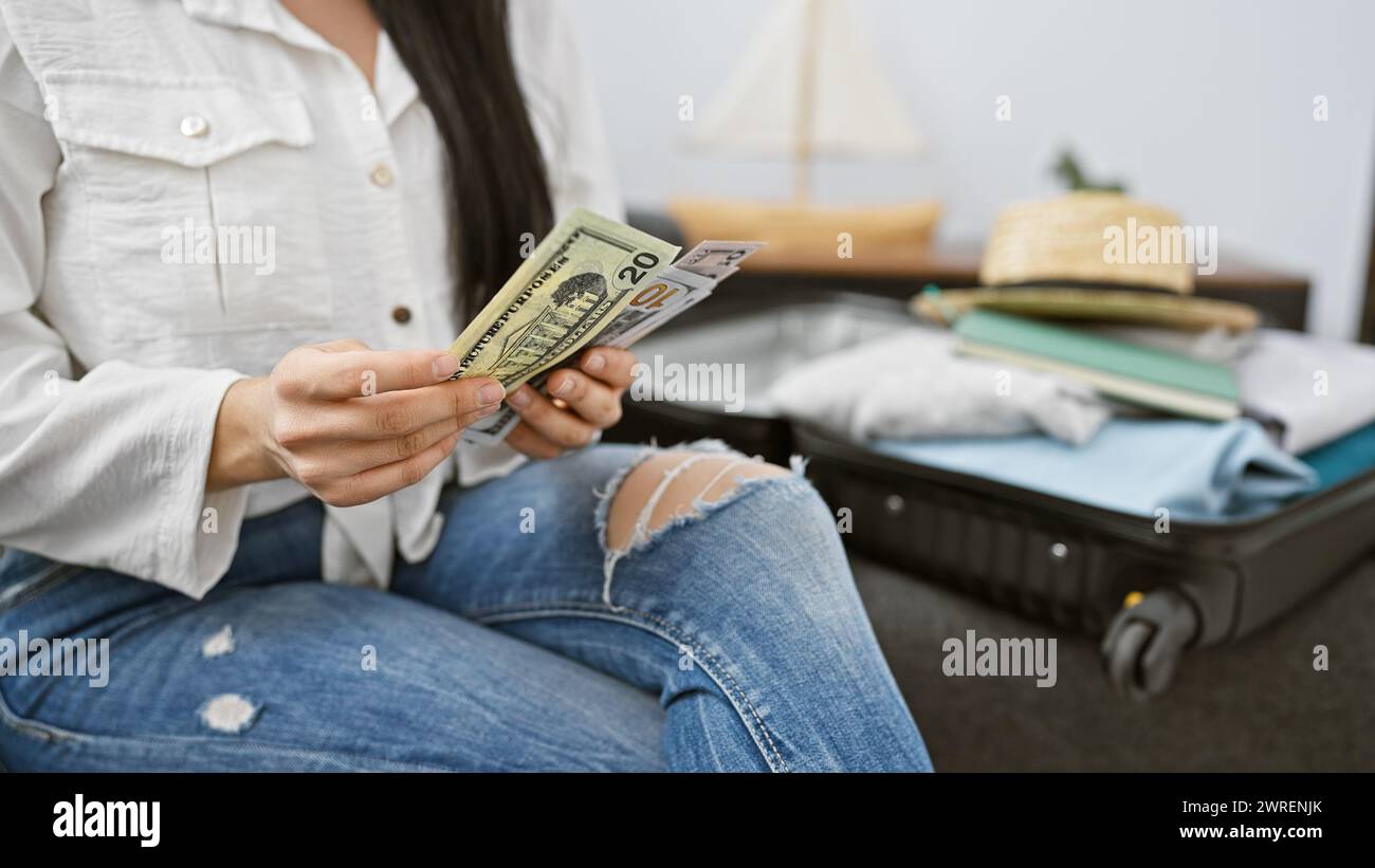 Une jeune femme hispanique nous compte des dollars avant de voyager, assise à l'intérieur avec une valise. Banque D'Images