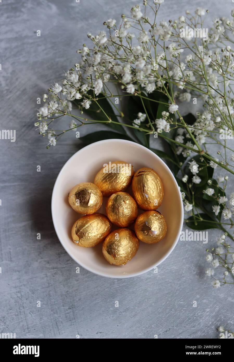 Œuf de Pâques en chocolat enveloppé dans une feuille d'or. Fleurs de printemps blanches et bonbons ester vue du dessus photo. Nature morte printanière brillante. Banque D'Images