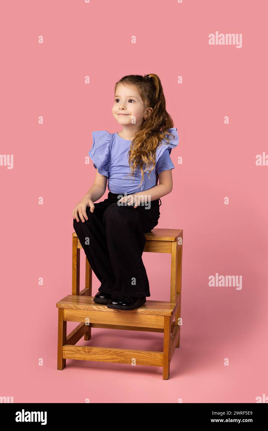 Une petite fille est assise sur une chaise et est grosse, la chaise est faite par son père charpentier. Elle est habillée d'une blouse bleue très élégante. Elle a l'air AWA Banque D'Images
