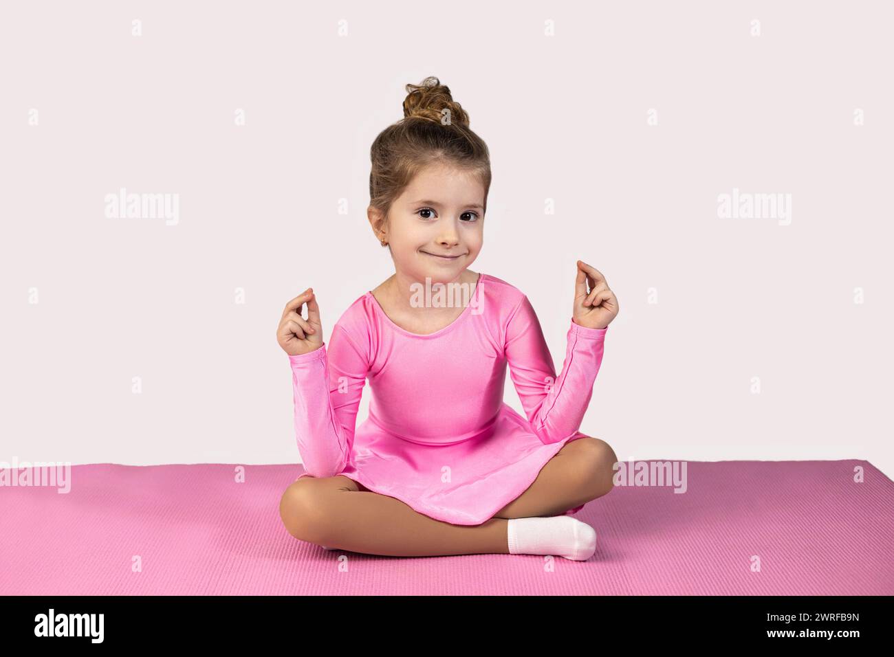 Petite fille gymnaste en position de lotus souriant magnifiquement à la méditation de caméra, assise sur un tapis de fitness rose portant une robe de gymnastique rose. Haute qualité Banque D'Images