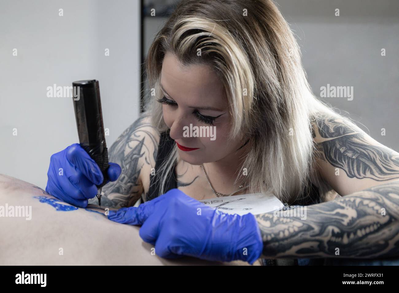 Photo horizontale une tatoueuse dévouée avec des tatouages à manches pleines applique son art, gravant un dessin sur le corps d'un client dans son studio. Concept busine Banque D'Images