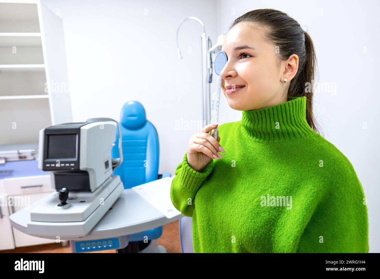 Une femme heureuse dans un pull vert se tient debout devant une machine d'examen de la vue, souriant tandis qu'elle fait des gestes vers l'équipement de bureau sur le bureau Banque D'Images