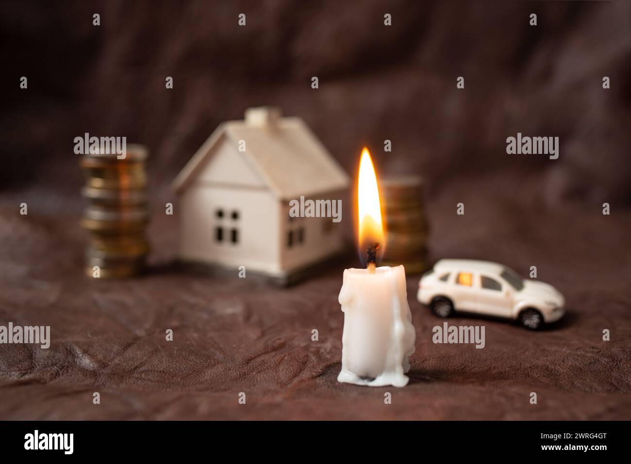 bougie brûlant avec des pièces de monnaie, maison miniature et voiture en arrière-plan, sur cuir brun, richesse face à la métaphore de la mort Banque D'Images