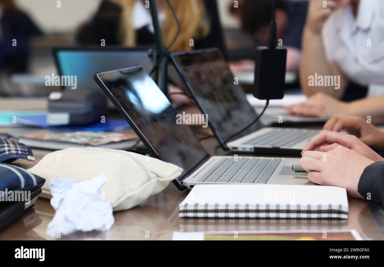 Élèves occupés travaillant à une table sur des ordinateurs portables avec des accessoires de cours typiques tels que des chargeurs, des livres, des stylos, des poubelles et des trousses à crayons. Banque D'Images