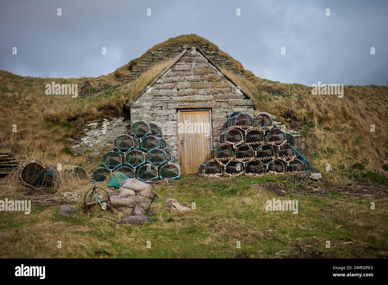 Chalet en pierre / hangar à l'entrée du port de Sinclairs Bay avec des casiers de homard et de crabe empilés à l'extérieur par une journée ensoleillée en Écosse rurale, prêt pour la pêche Banque D'Images