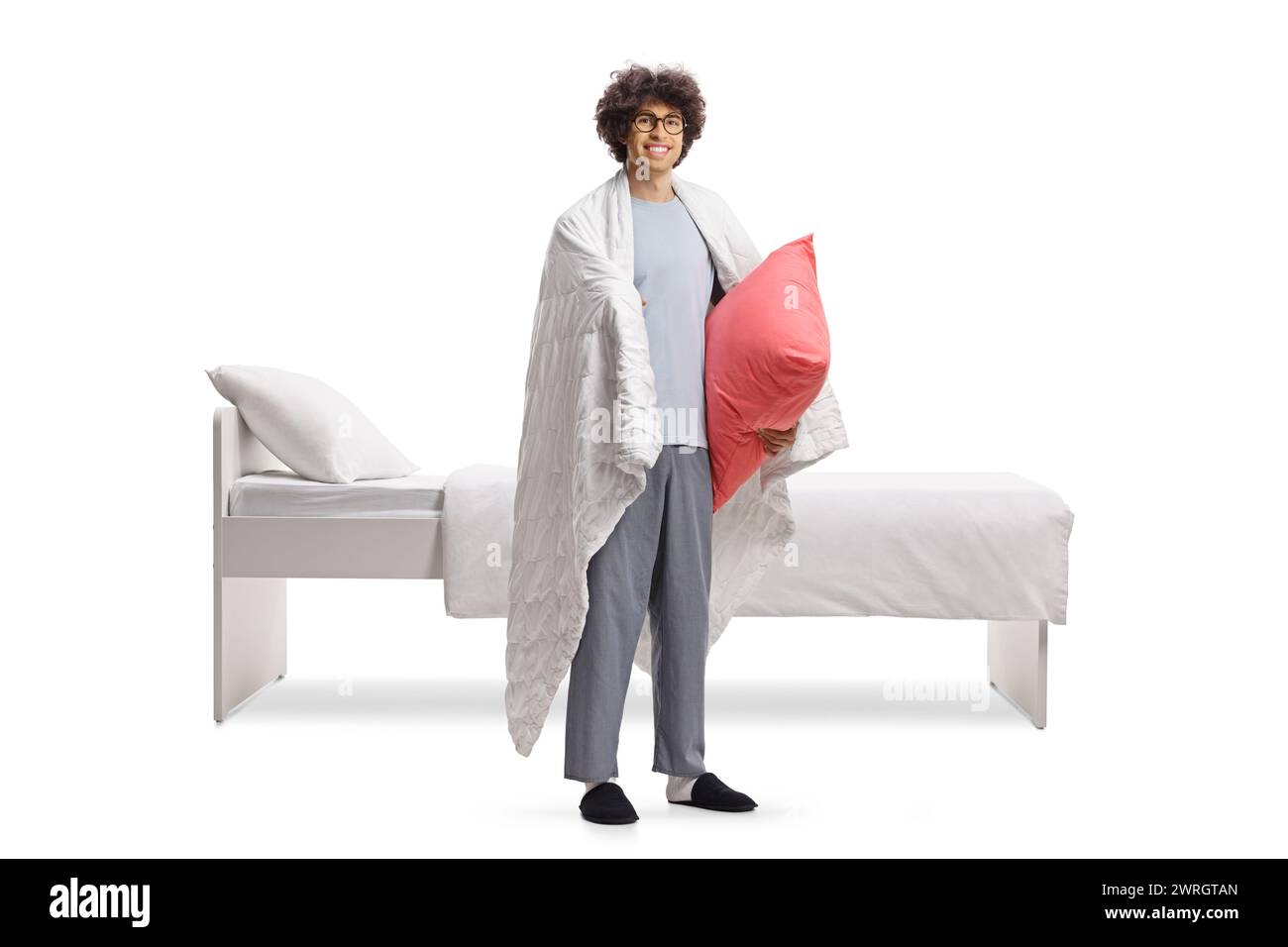 Jeune homme en pyjama enveloppé dans une couverture debout à côté d'un lit et tenant un oreiller isolé sur fond blanc Banque D'Images