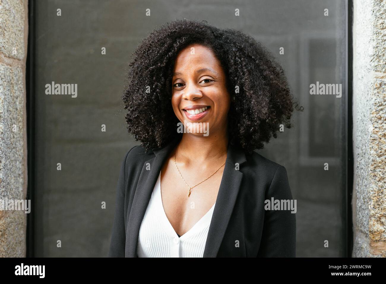 Une femme afro-américaine joyeuse en tenue d'affaires pose avec le sourire à l'extérieur d'un immeuble de bureaux, exsudant la confiance professionnelle. Banque D'Images