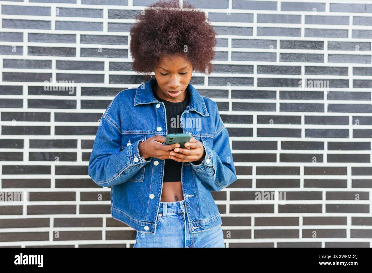 Une jeune femme afro-américaine se concentre intensément sur son smartphone, debout devant un mur de briques noires et blanches frappant Banque D'Images