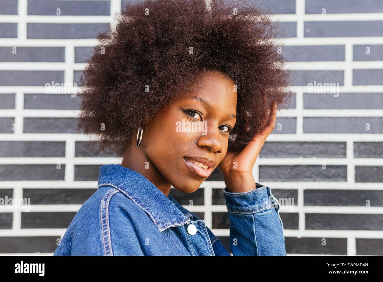 Femme afro-américaine élégante donne un regard de côté confiant, ses cheveux naturels contrastant avec le mur de briques urbain graphique derrière elle Banque D'Images