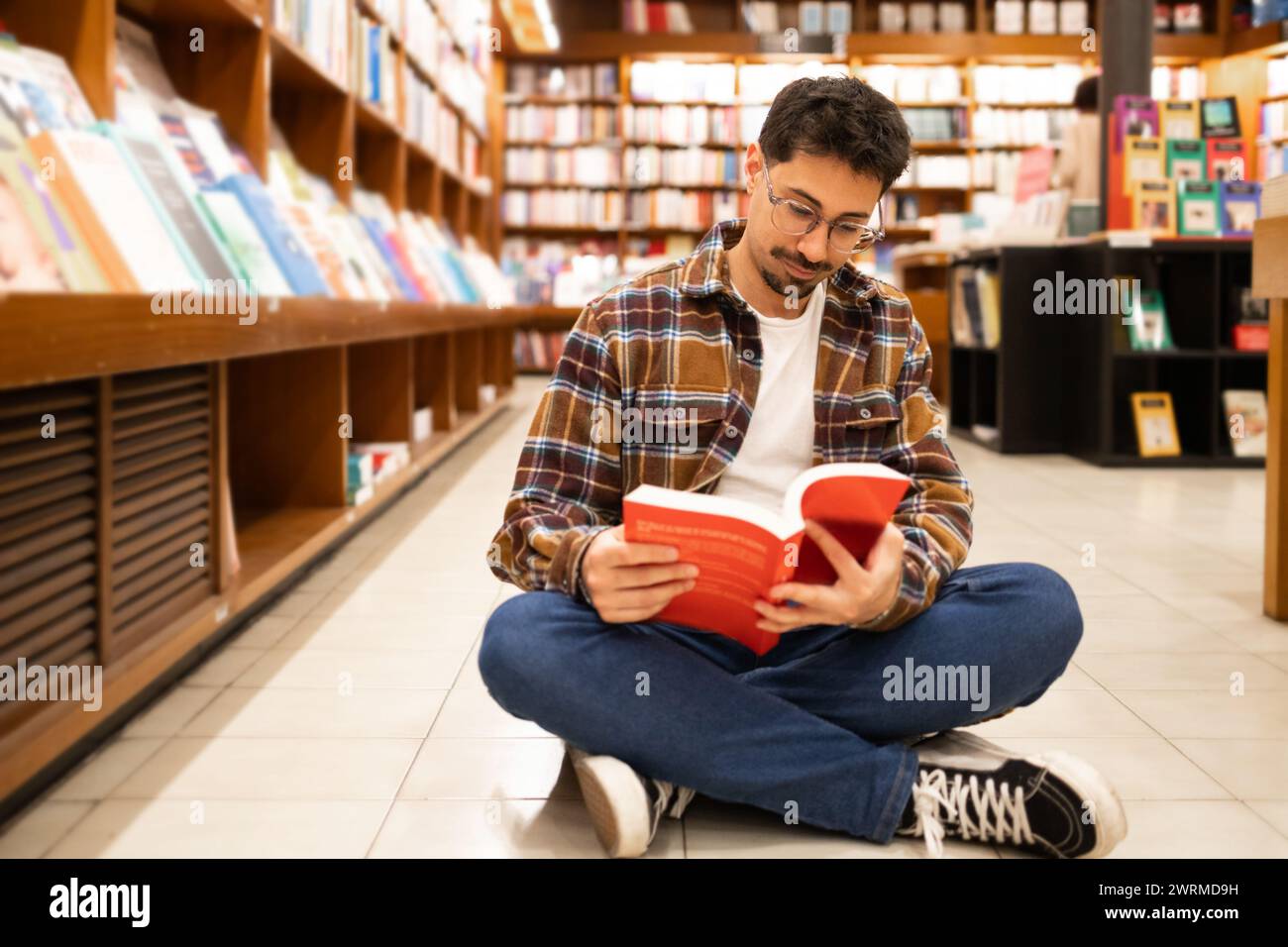 Un jeune homme est assis les jambes croisées sur le sol d'une librairie, engrossé dans un livre rouge, entouré d'étagères pleines de livres. Banque D'Images