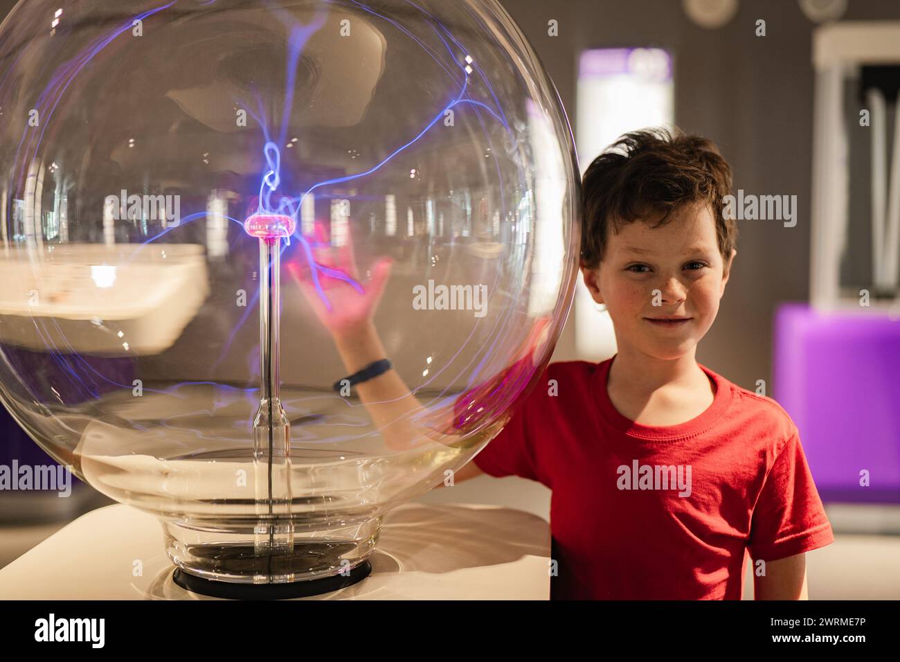 Un garçon curieux dans une chemise rouge semble étonné par une exposition de globe à plasma dans un musée des sciences, sa main interagissant avec l'électricité statique. Banque D'Images