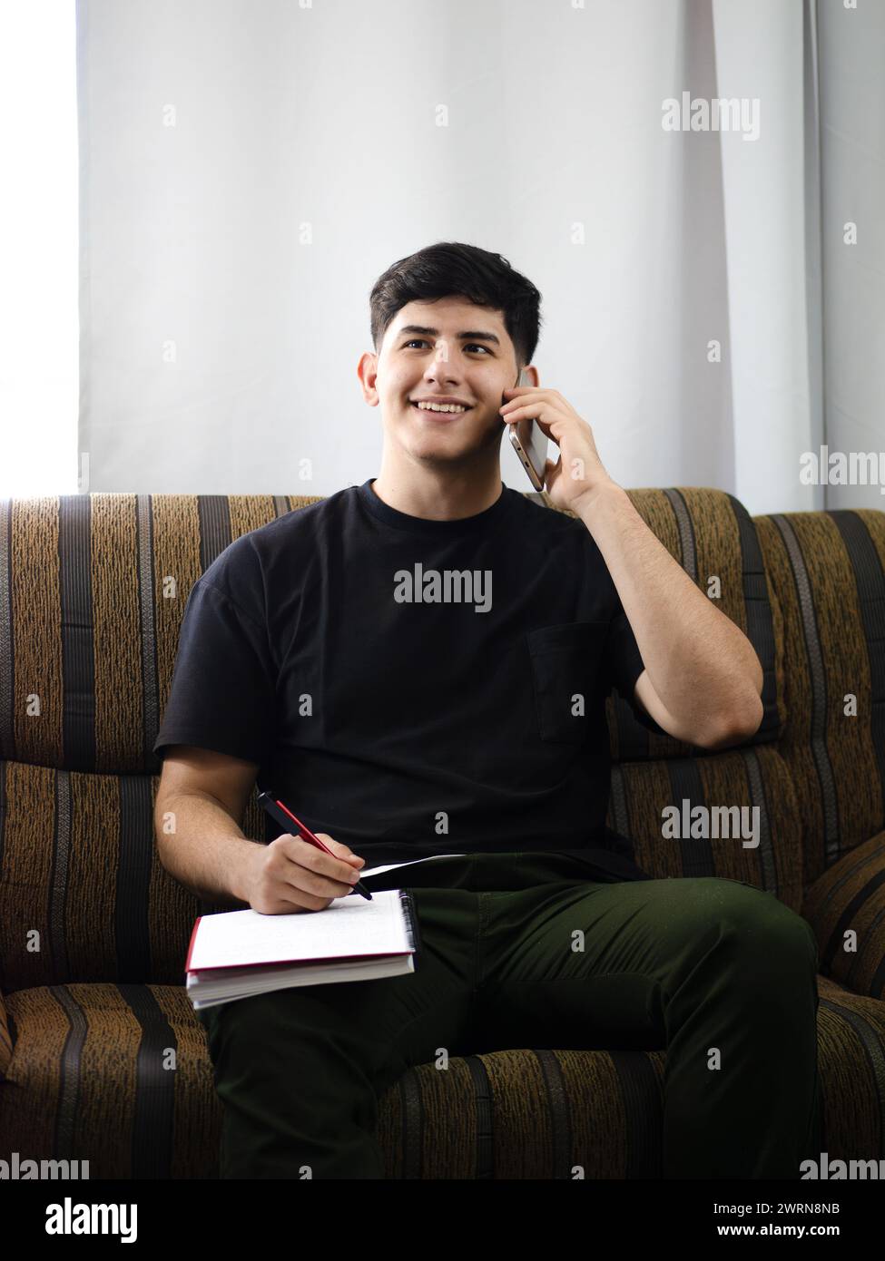 Un jeune homme souriant parle au téléphone tout en écrivant dans un ordinateur portable, confortablement assis dans un bureau ou à la maison. Travail à distance, organisation personnelle Banque D'Images