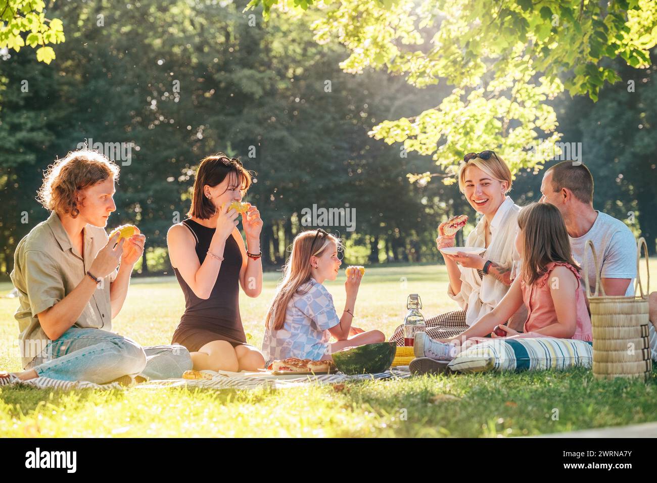 Grande famille assis sur la couverture de pique-nique dans le parc de la ville pendant le week-end dimanche journée ensoleillée. Ils sourient, rient et mangent du maïs bouilli et de la pastèque Banque D'Images