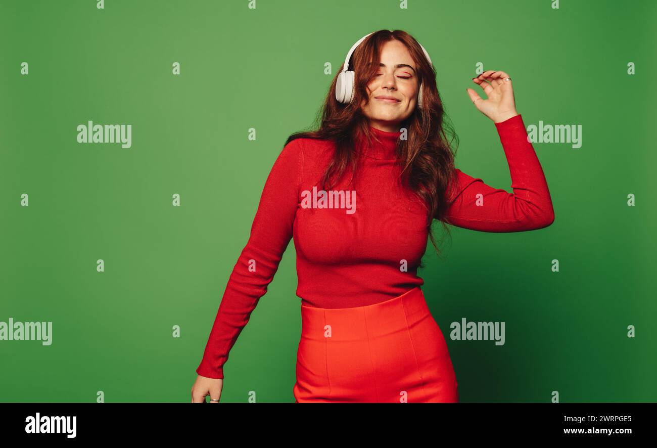 Jeune femme en pull décontracté se tient sur un fond vert écoutant de la musique à travers des écouteurs sans fil. Elle bouge et danse au rythme d'une che Banque D'Images