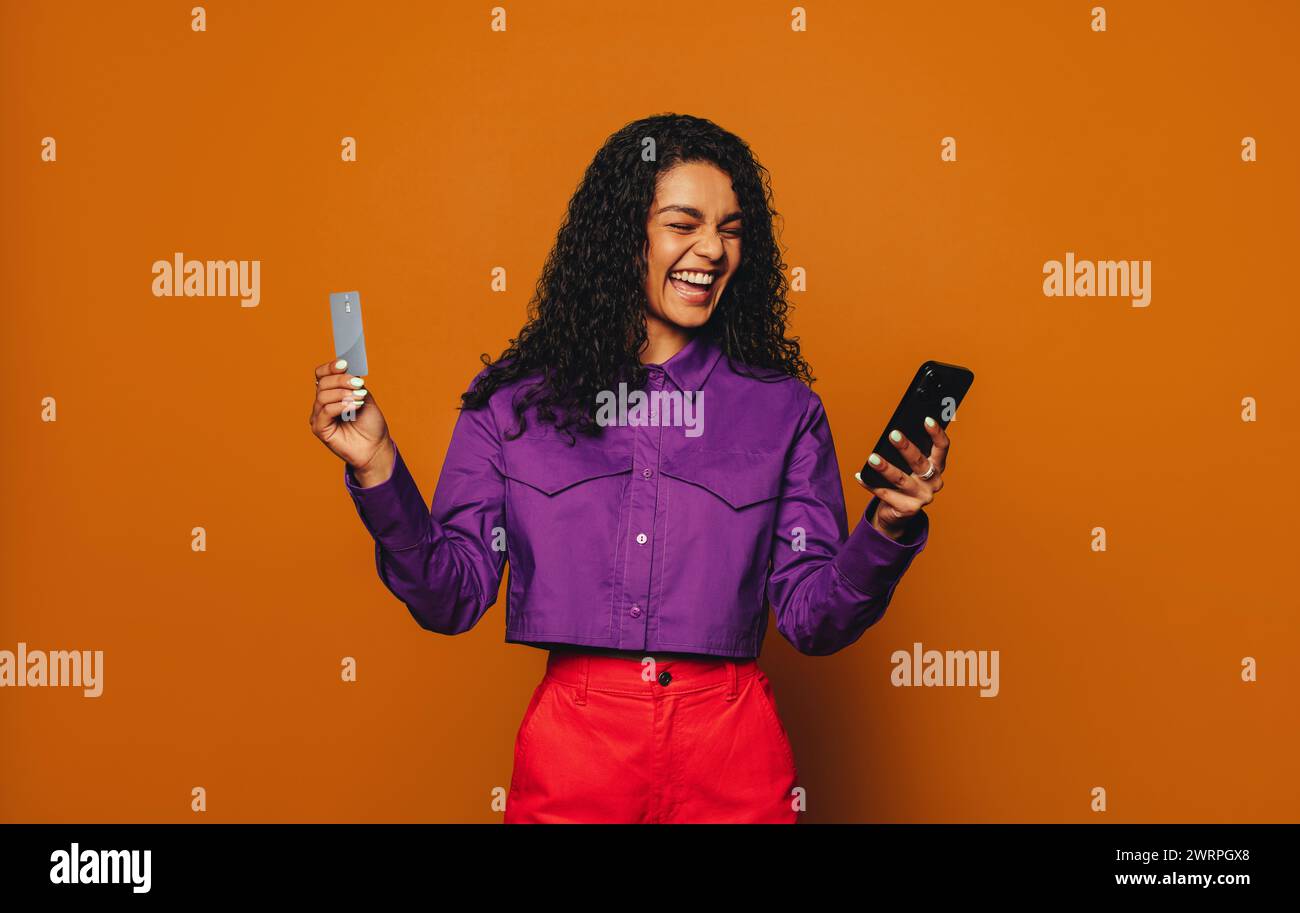 Femme joyeuse avec les cheveux bouclés se tient sur un fond orange vibrant. Elle sourit tout en payant ses achats en ligne sur son téléphone portable en utilisant un crédit c. Banque D'Images