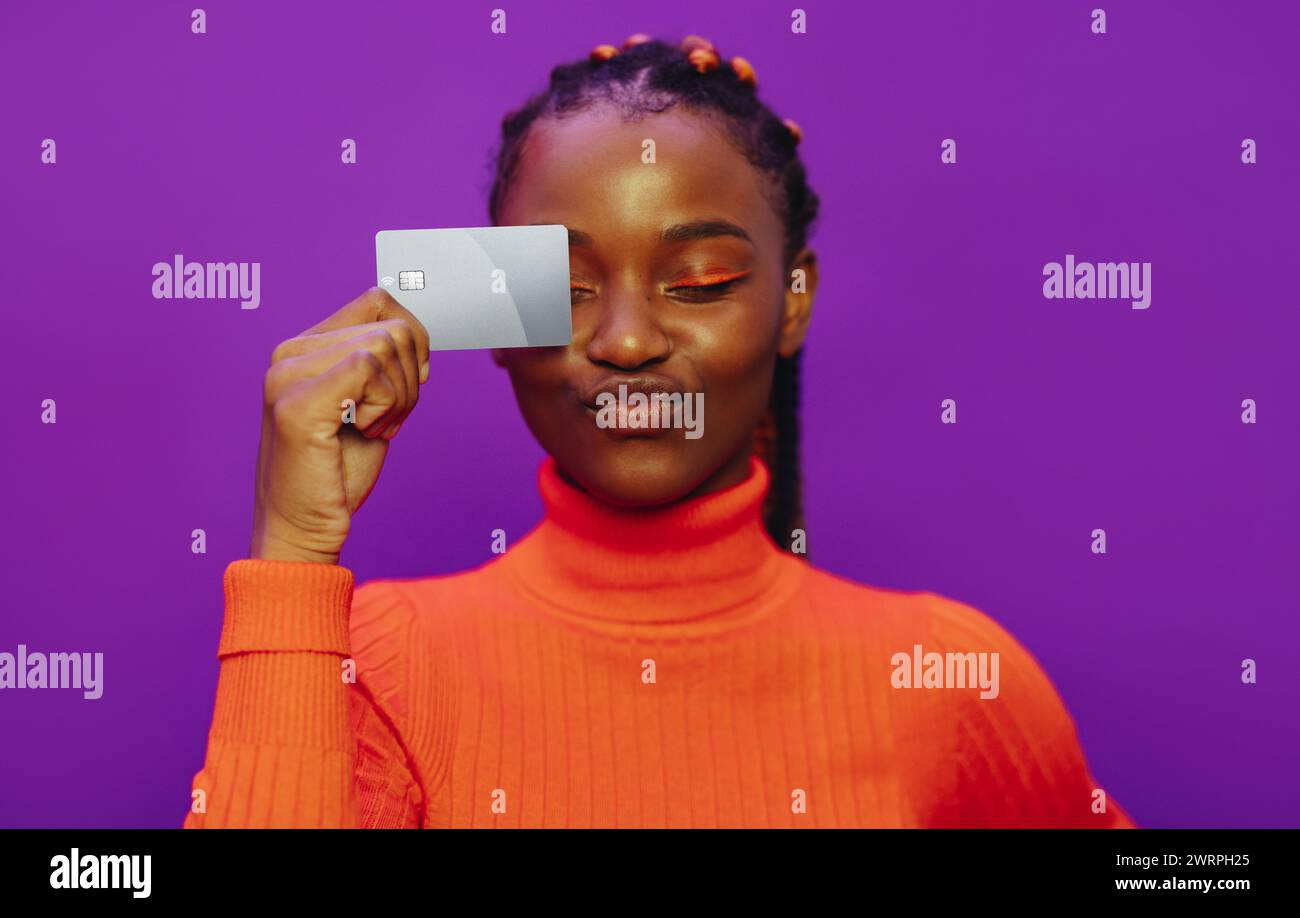 Femme africaine dans des vêtements vibrants et décontractés se dresse sur un fond violet. Les yeux fermés, elle tient une carte bancaire, payant électroniquement avec un robinet... Banque D'Images