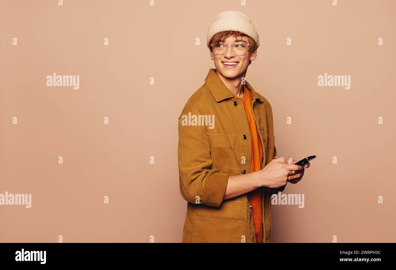 Joyeux homme se tient dans un studio portant des vêtements décontractés et une veste en denim. Tenant un téléphone portable, il communique avec un sourire éclatant. La pêche BA Banque D'Images