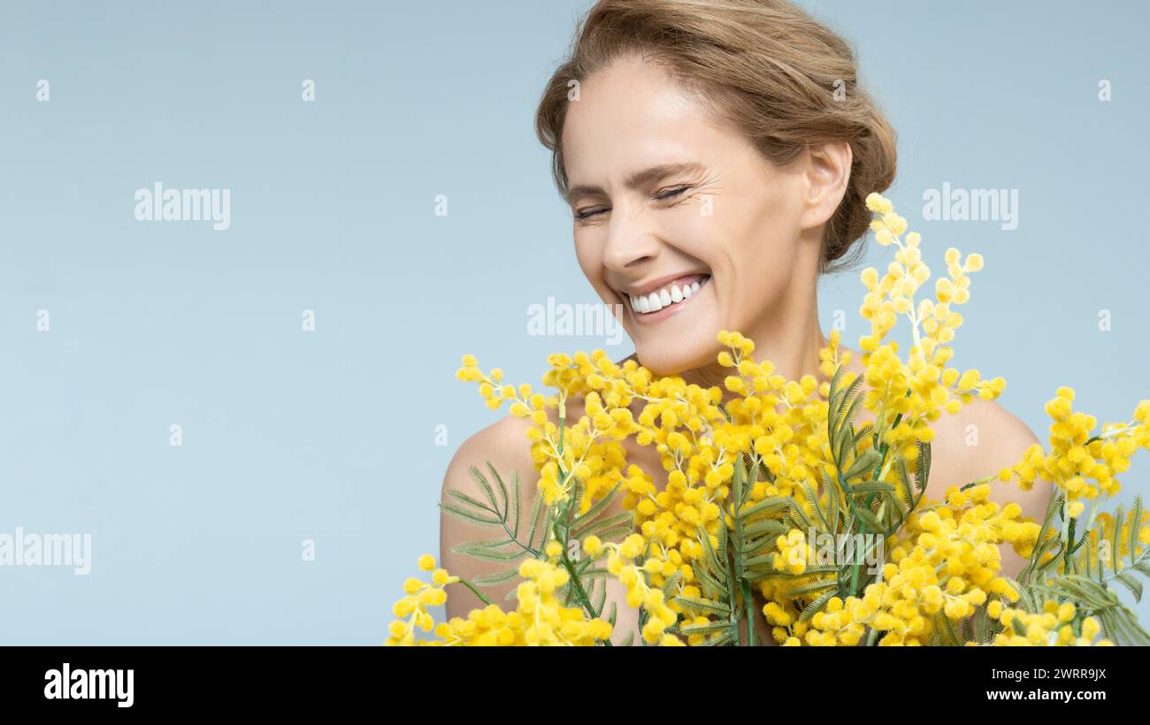 Portrait heureux de femme mature confiante et en bonne santé avec un beau bouquet de mimosa sur fond bleu. Sourire radieux de belle femme Banque D'Images