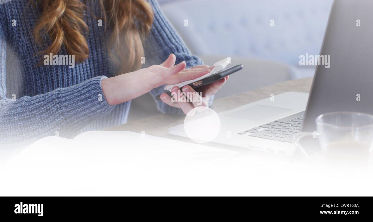 Image de taches blanches sur une femme caucasienne essuyant un smartphone à l'aide d'un ordinateur portable à la maison Banque D'Images