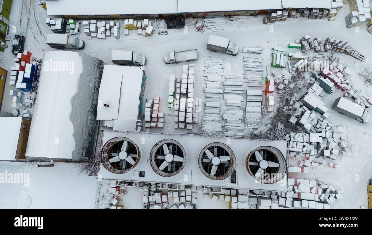 Photographie par drone de ventilateurs de refroidissement à l'échelle industrielle et de matériaux de construction dans un entrepôt en plein air couvert par la neige pendant la journée nuageuse d'hiver Banque D'Images