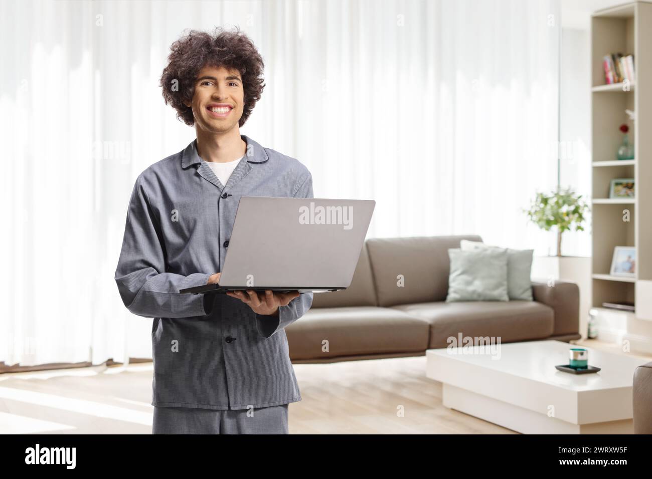 Jeune homme en pyjama debout dans un salon avec un ordinateur portable et souriant à la caméra Banque D'Images