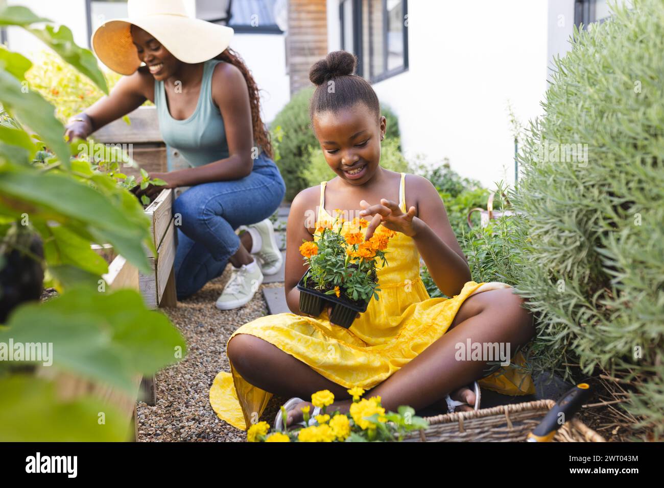 Une jeune fille afro-américaine admire une fleur en pot dans le jardin à la maison, avec sa mère derrière Banque D'Images