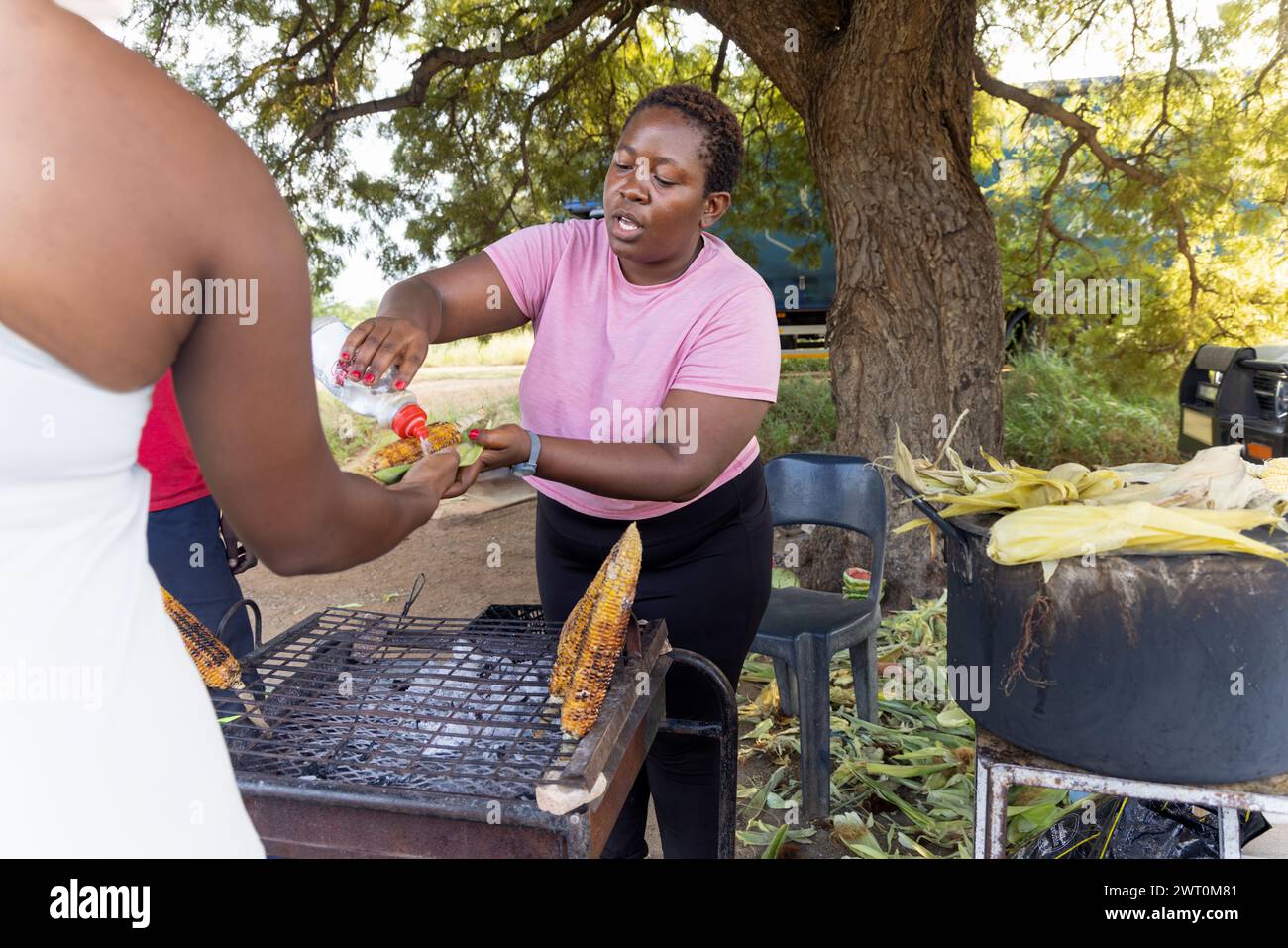 femme africaine vendeuse de rue vendant du maïs grillé à un client dans une rue de la ville sur le trottoir, pot bouillant dans le fond Banque D'Images