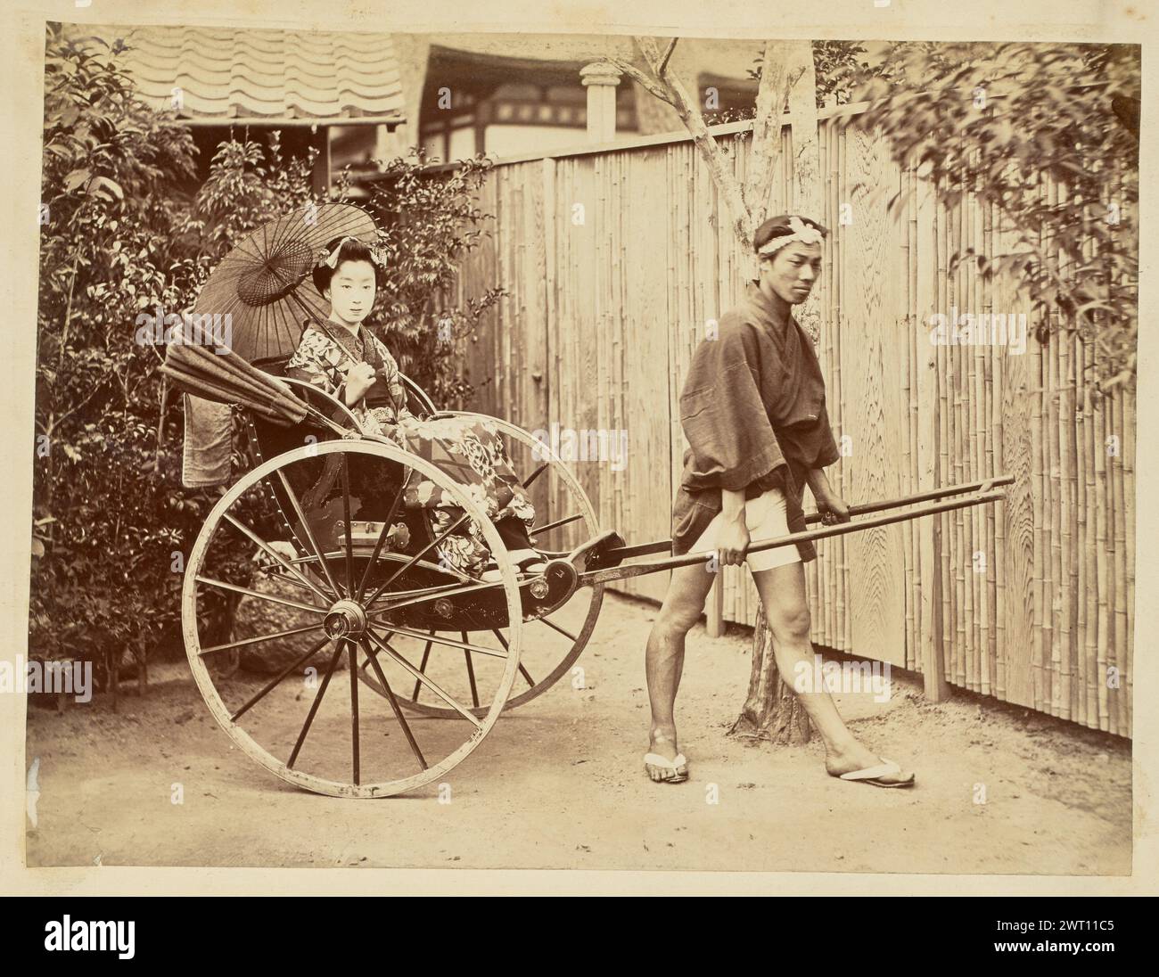 Femme japonaise dans un pousse-pousse. Inconnu, photographe des années 1850–1890 vue d'un homme debout entre les pôles d'un pousse-pousse, les tenant debout avec une jambe tendue devant lui et son pied doucement pointé. Une femme est assise dans le pousse-pousse, portant une robe japonaise traditionnelle, et tenant un parasol ouvert, ou parapluie en papier huile, par-dessus son épaule pendant qu'elle regarde l'appareil photo. Banque D'Images