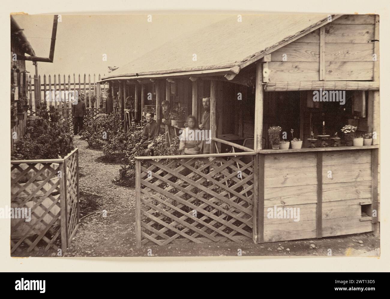 Camp militaire. Charles Parker, photographe (britannique, actif dans les années 1860) vers 1864 vue d'une caserne militaire en bois, séparée du bâtiment suivant par un petit sentier bordé de plantes cultivées. Sont également visibles un certain nombre d'hommes caucasiens vêtus d'uniformes militaires et un garçon japonais vêtu de vêtements traditionnels. Banque D'Images