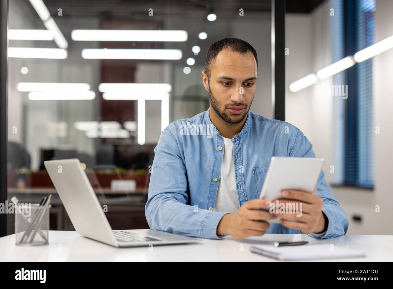 Un homme sérieux d'Amérique latine en chemise bleue travaille dans un bureau moderne, s'assoit à un bureau avec un ordinateur portable et se concentre sur l'utilisation d'une tablette dans ses mains. Banque D'Images