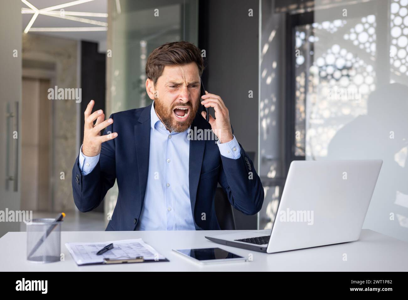 Jeune homme d'affaires masculin en colère assis à un bureau dans le bureau et parlant agressivement au téléphone, criant et faisant des gestes avec ses mains. Banque D'Images