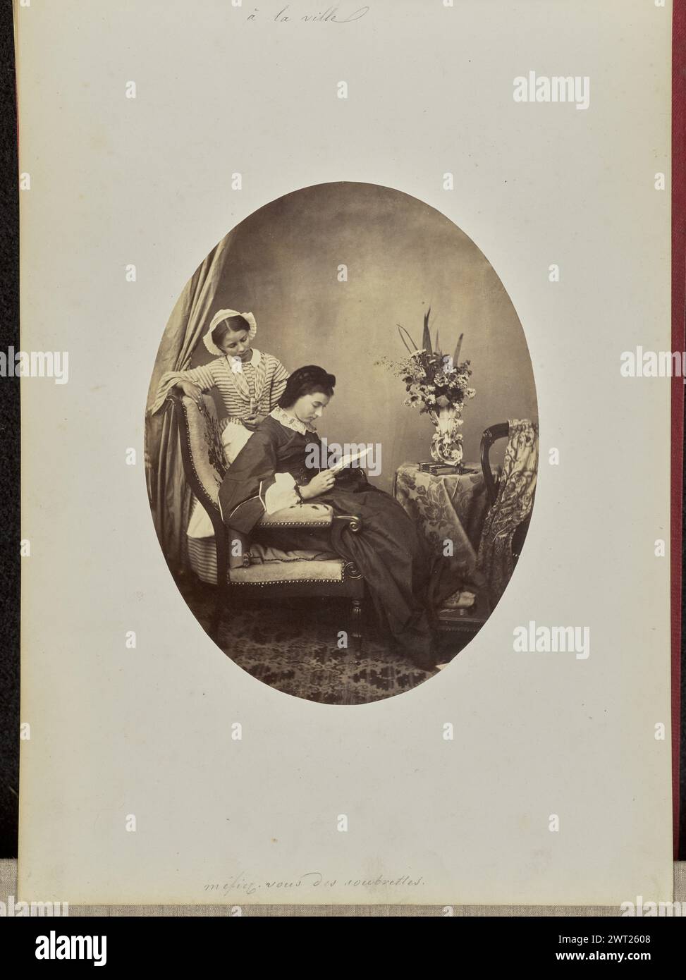 á la ville, méfiez-vous des soubrettes. Comte de Charly, photographe (actif des années 1850 - 1880) 1862 Une femme assise sur une chaise, penchée sur un morceau de broderie. Une deuxième femme portant une robe rayée se tient derrière elle. La femme dans la robe rayée et le bonnet repose une main sur le dos de la chaise et semble regarder par-dessus l'épaule de la femme assise. (Recto, monture) haut centre, encre noire : 'à la ville' ; bas centre, encre noire : 'méfiez vous des soubrettes'; Banque D'Images
