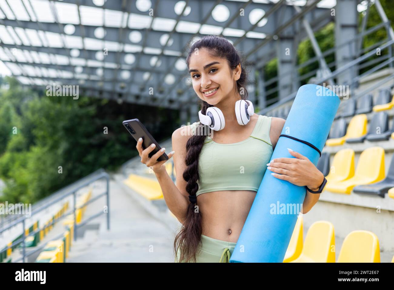 Portrait de jeune femme indienne sportive debout sur la tribune du stade en costume et écouteurs, tenant tapis d'exercice et téléphone, souriant à la caméra. Banque D'Images