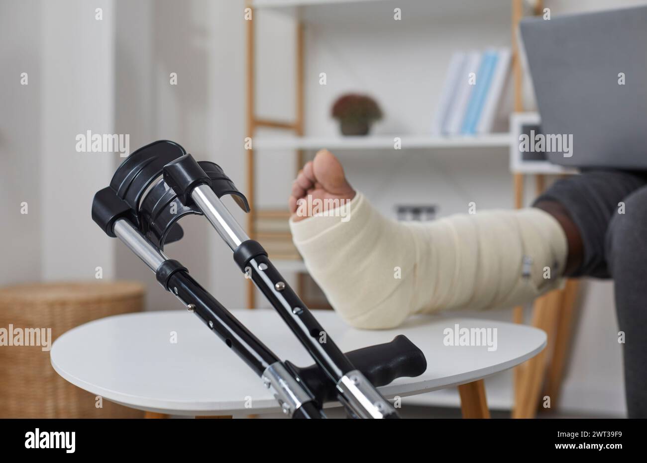 Femme avec une jambe cassée utilisant son ordinateur portable, le pied reposant sur un tabouret, avec des béquilles à côté Banque D'Images