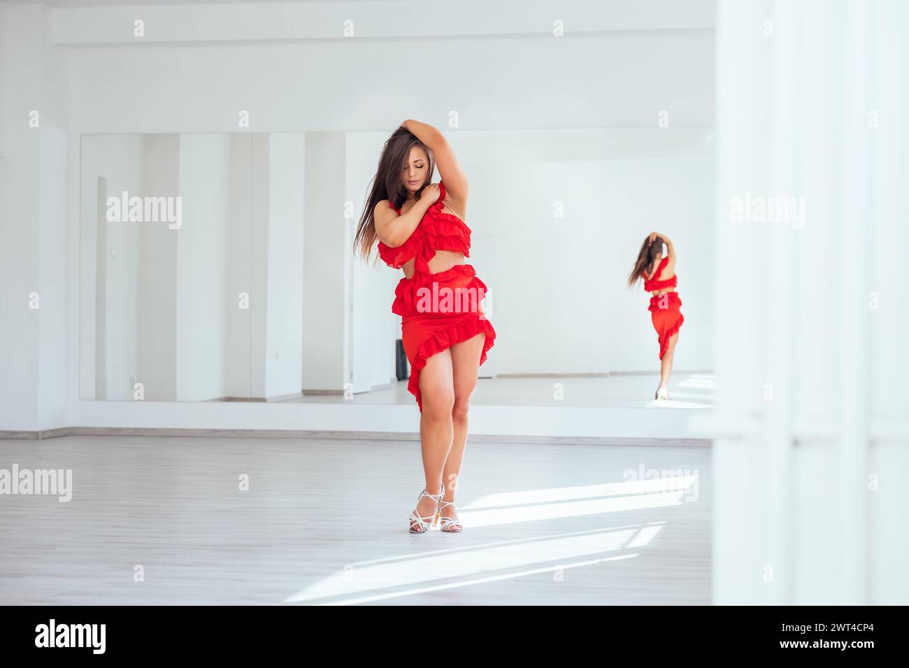 Femme gracieuse habillée en robe de danse latine rouge faisant des pas de danse élégants dans la grande salle de couleur blanche avec grand mur miroir. Expressions des gens pendant da Banque D'Images