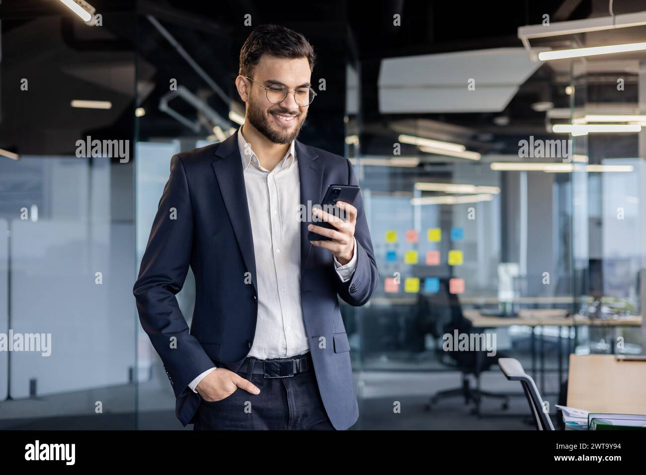 Un jeune homme d'affaires joyeux en costume interagit avec son smartphone dans un cadre de bureau lumineux et contemporain. Banque D'Images