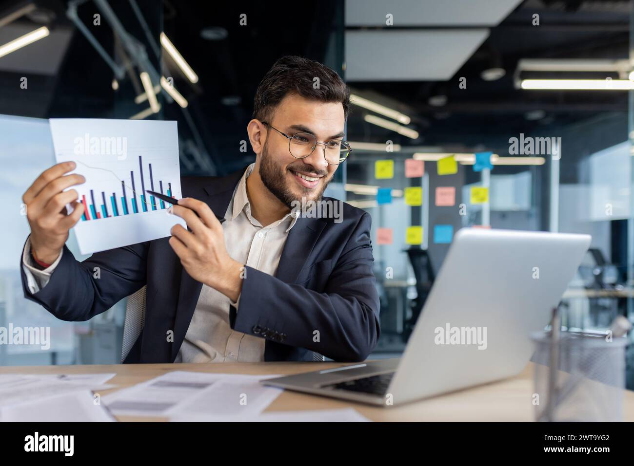 Un homme d'affaires souriant dans un costume affichant un graphique à barres à la caméra, illustrant la croissance et le succès de l'entreprise dans un cadre de bureau moderne. Banque D'Images