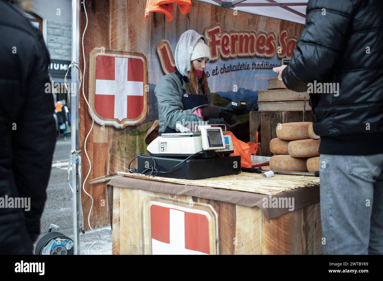 Lanslevillard, France, 10 janvier 2024 : une vendeuse à capuche debout derrière le stand avec du fromage sert un client sur le marché de la ville Banque D'Images