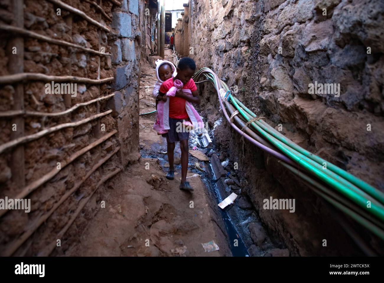 Une jeune fille passe devant les couloirs dans le bidonville de Kibera en transportant son frère cadet. Kibera, le plus grand bidonville de Nairobi et d'Afrique, abrite plus de Tha Banque D'Images