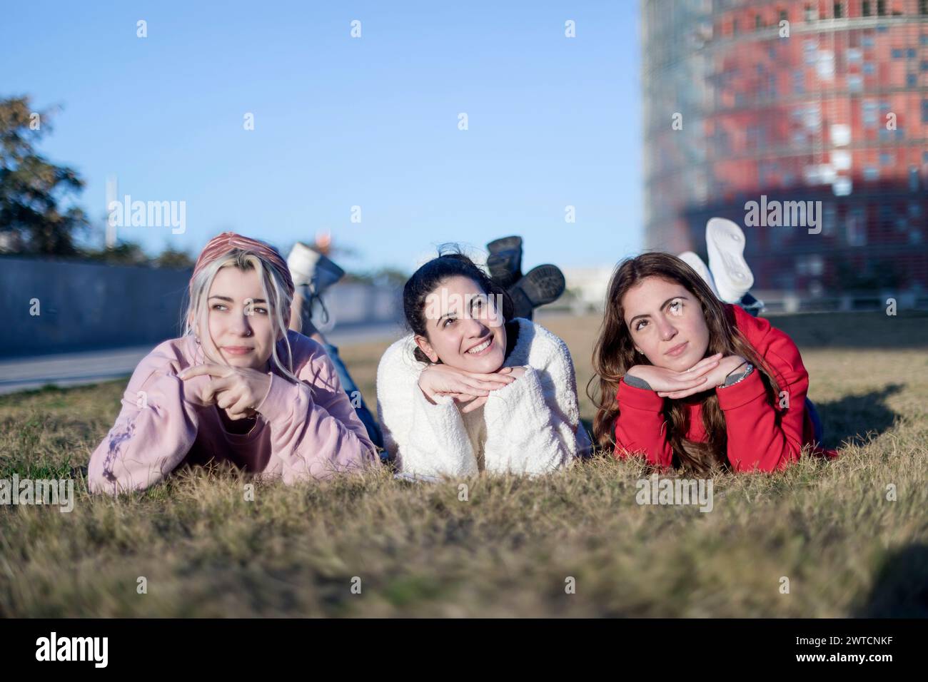 Trois jeunes femmes allongées sur l'estomac sur l'herbe, posant de manière ludique les mains sous le menton, avec un ciel clair et un fond urbain Banque D'Images