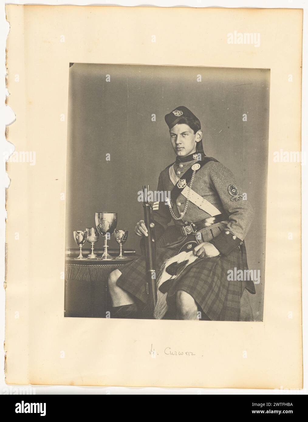 H. Curwen. Probablement Robert Ewing Curwen, photographe (britannique, vers 1841 - 1934) vers 1895 Un portrait de Henry Curwen, un des fils de Robert Ewing Curwen. Il est vu en tenue militaire, portant un kilt et un glengarry. Il tient une arme d'une main et il y a un plateau avec des gobelets à côté de lui. (Recto, monture) centre inférieur, crayon : 'H. Curwen' ; (verso, monture) haut droit, crayon : '27'; Banque D'Images