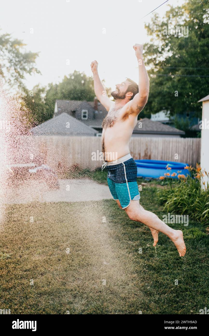 L'homme souriant saute à travers la pulvérisation d'eau de tuyau à l'extérieur pendant l'été Banque D'Images