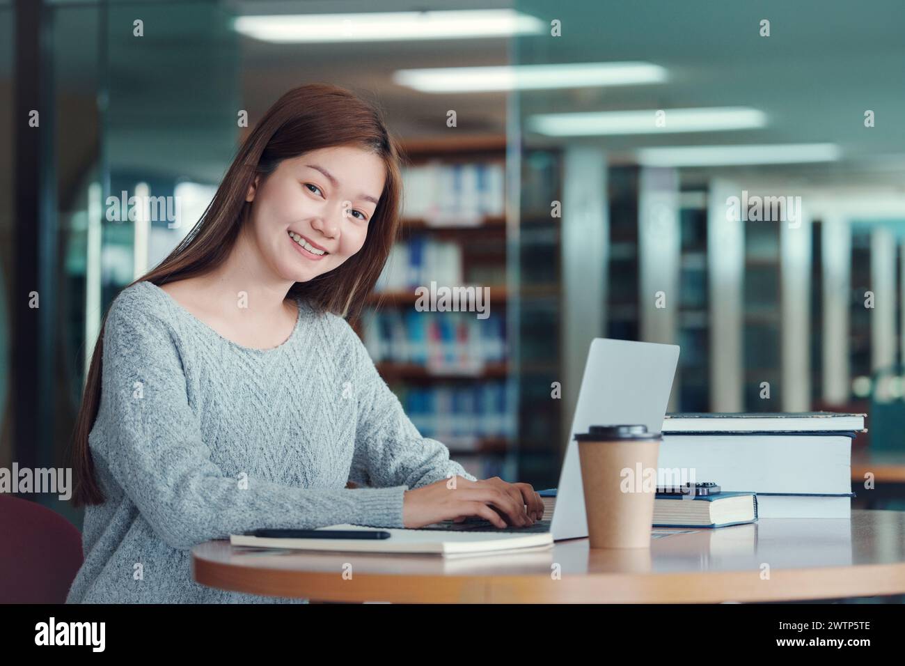 Heureuse jeune étudiante étudiante à la bibliothèque du collège, assise au bureau, utilisant un ordinateur portable. Banque D'Images
