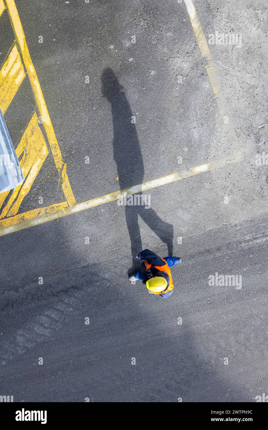 Un travailleur de la construction portant un casque de sécurité marche sur le tarmac. Il projette une longue ombre. Stuttgart, Bade-Wuerttemberg, Allemagne Banque D'Images