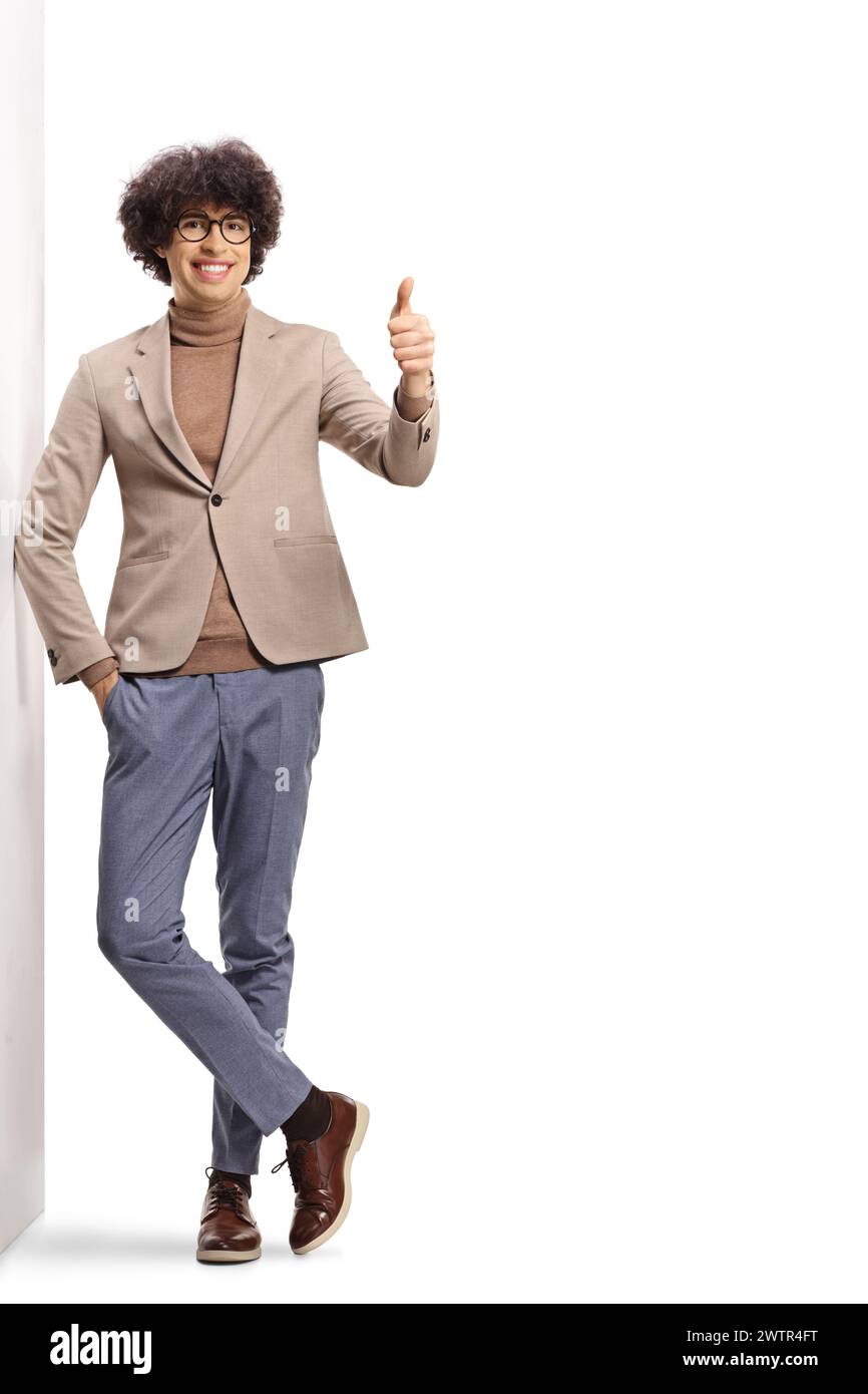 Portrait en pied d'un jeune homme joyeux avec des lunettes appuyées sur un mur et faisant des gestes pouces isolés sur fond blanc Banque D'Images