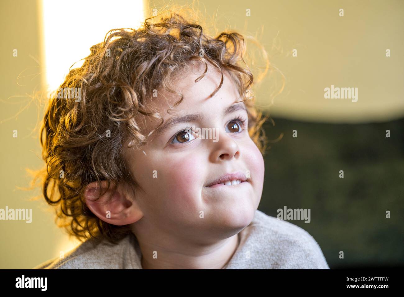 Jeune enfant avec des cheveux bouclés regardant vers le haut réfléchi dans une pièce chaleureusement éclairée. Banque D'Images