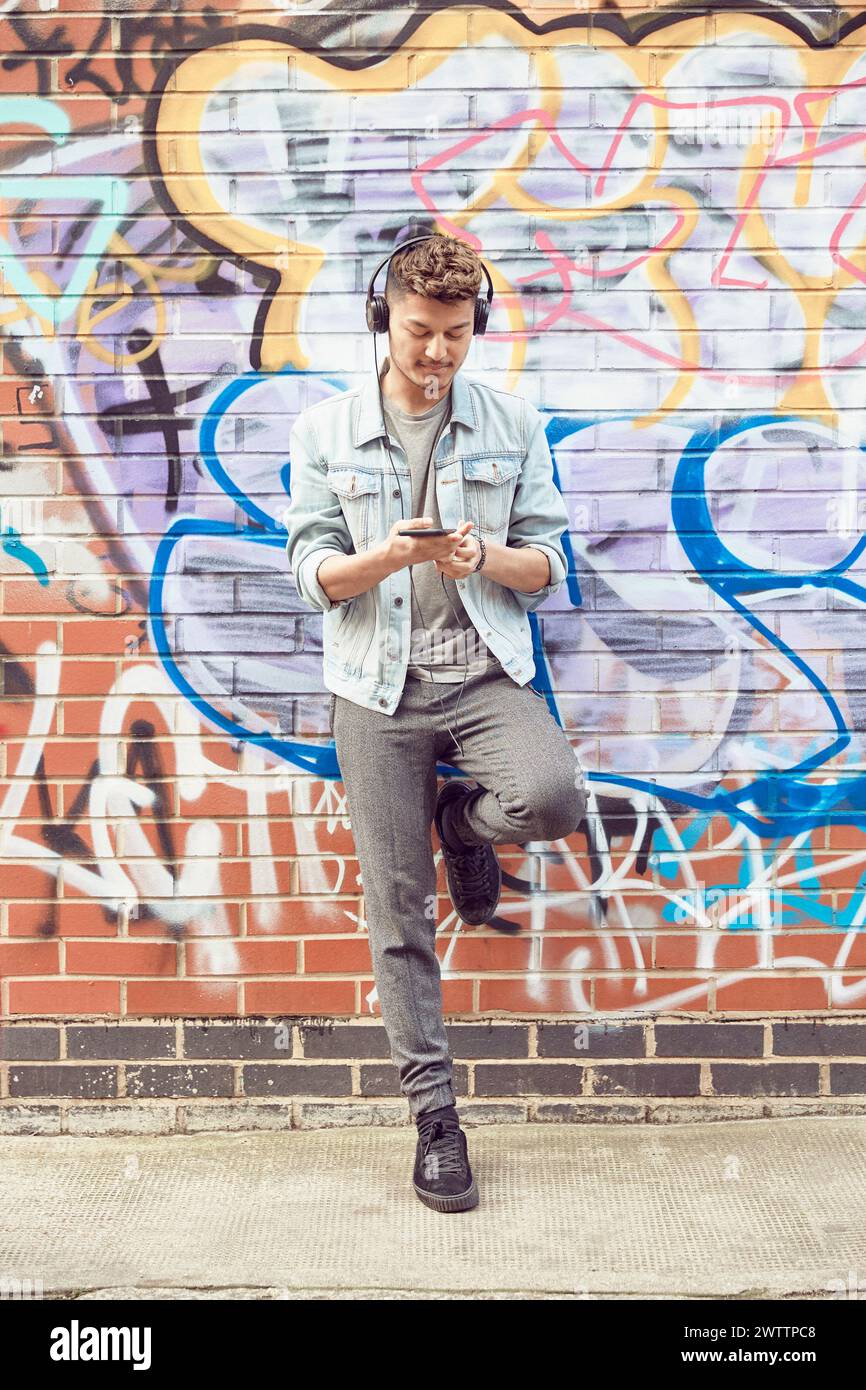 Homme avec des écouteurs vérifiant le téléphone par le mur de graffiti Banque D'Images